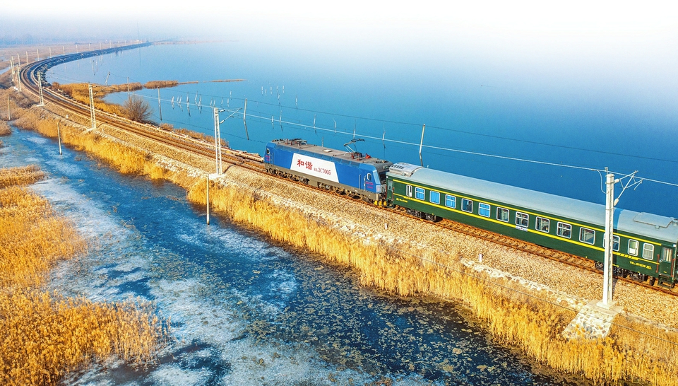 茹雅摄一列长长的火车奔驰在水上,一侧是高耸山峦,一侧是湛蓝湖面.